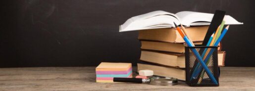 A stack of school supplies, including text books, pencils, and sticky notes sits on a desk askew.