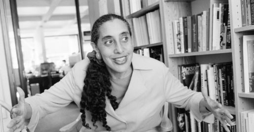 A black and white photo of a young women with long hair tied back; she opens her arms in excitement and sits in front of shelves and shelves of books.