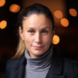A white woman stares warmly out at the camera, wearing a turtle neck and a blazer. Behind her lights flash in the distance.