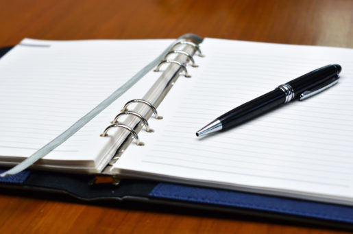 A note book on a table lies splayed open to a blank page with a pen lying across it askew.