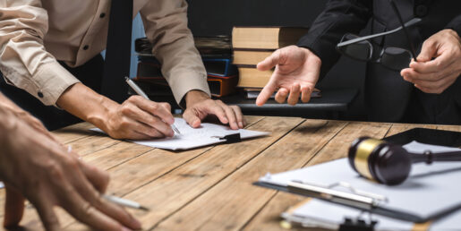 Image that shows the hands of a few individuals leaning over a table and holding pens ready to sign documents that are laid out in front of them.