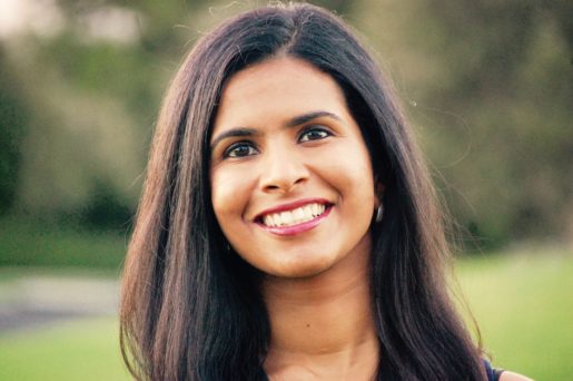 A south Asian woman with long brown hair smiles warmly against a blurred green backdrop.
