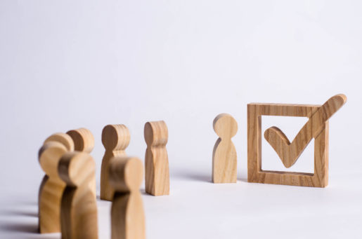 Wood figurines march toward a wooden checkmark, illustrating people going to vote.