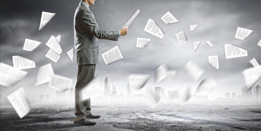 A businessman examines a document while papers fly around him in a barren landscape.