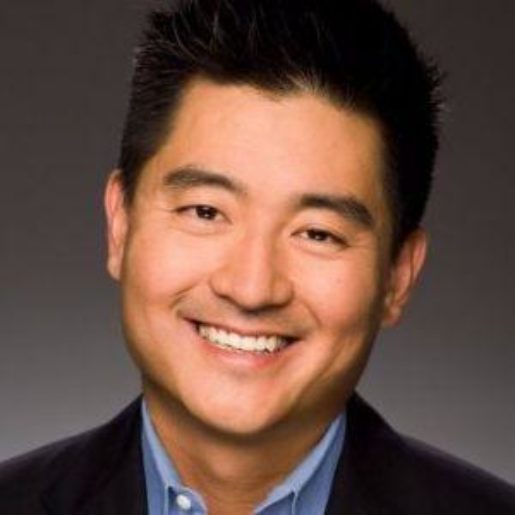 A man with dark hair wearing a suit jacket and a blue shirt smiles against a gray background.