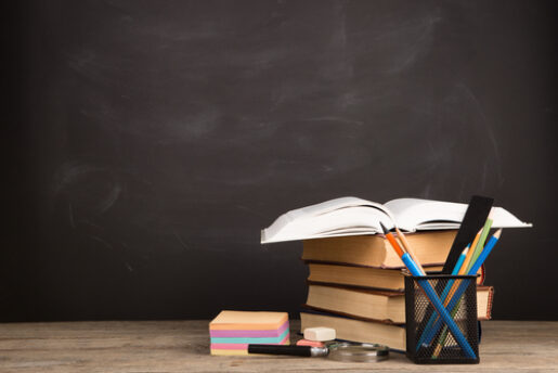 A stack of school supplies, including text books, pencils, and sticky notes sits on a desk askew.
