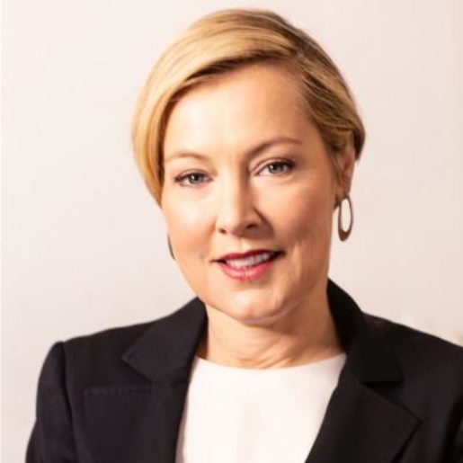 A white woman with short blond hair wearing a blazer smiles subtly against a pink background.