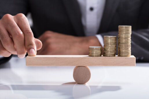 Many coins sit on a wooden balance beam while a hand holds the other side level.